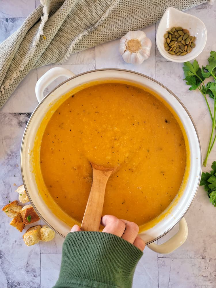 hand holding a wooden spoon in a pot of butternut squash, croutons in the background with a small ramekin with pumpkin seeds, fresh herbs and a whole garlic bulb