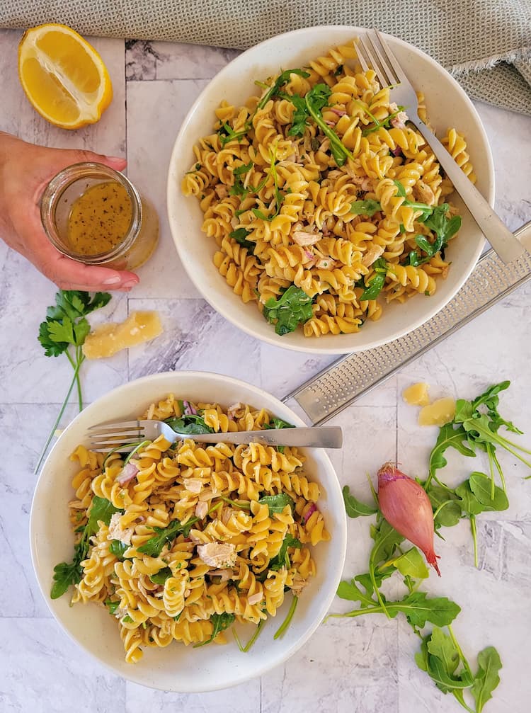 two bowls of pasta salad with tuna, arugula on the top and side with a shallot, a half a lemon and a hand holding a small jar of the dressing