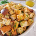 side view of a bowl of croutons, fresh herbs and a jar of olive oil in the background