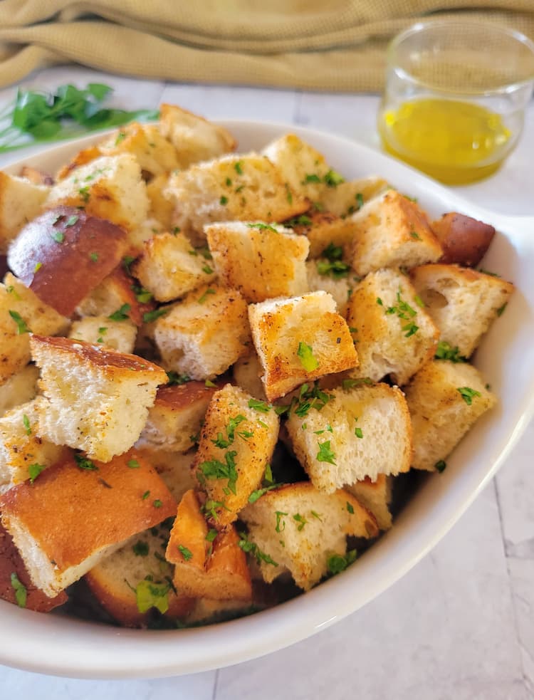 side view of a bowl of croutons, fresh herbs and a jar of olive oil in the background