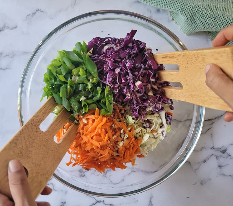 large bowl with chopped green onions, grated carrots, shredded red and green cabbage with dressing drizzled on top, hand with 2 large wooden forks about to mix it