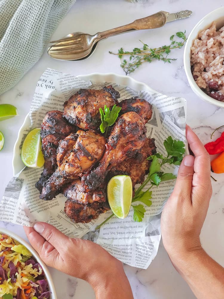 hands holding a bowl of jerk chicken garnished with parsley and lime wedges. Coleslaw, rice and peas, fresh thyme, lime and habaneros surrounding with spoons and fork