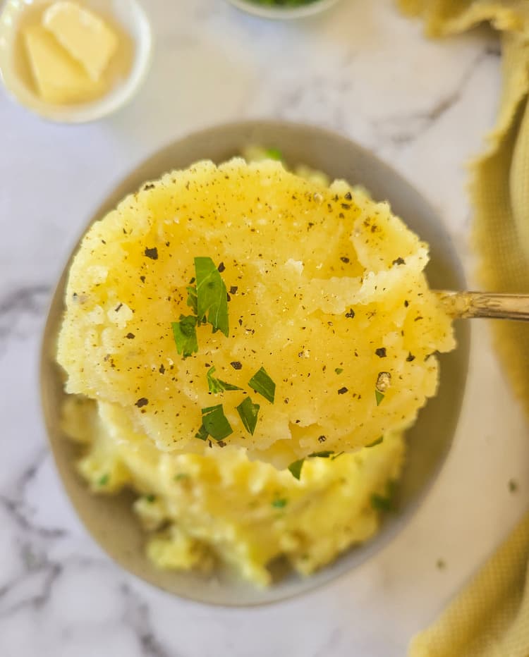 spoonful of mashed potatoes being held up over a bowl of the rest of them, garnished with fresh parsley, ramekin of butter in the background