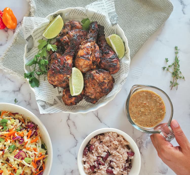newspaper lined plate with jerk chicken pieces garnished with fresh lime wedges and parsley, hand holding a pitcher of jerk sauce, bowl of rice and peas and coleslaw in the background with habaneros and fresh thyme sprigs