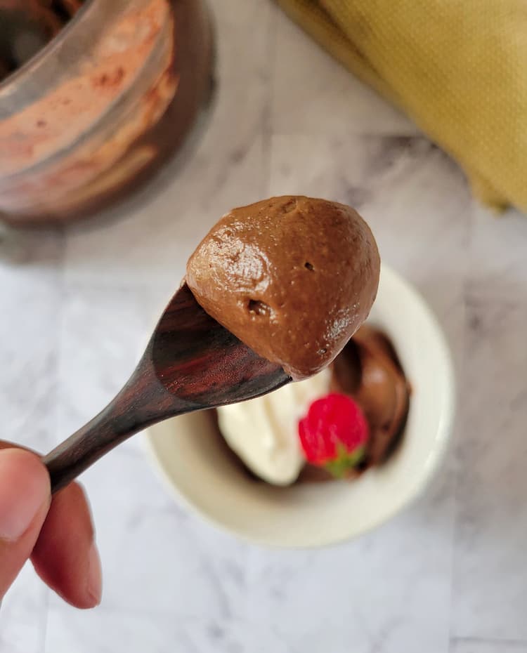 hand holding up a wooden spoon with some chocolate mousse with avocado on it over a bowl of the rest with whipped cream and a strawberry, food processor base with more mousse in the background