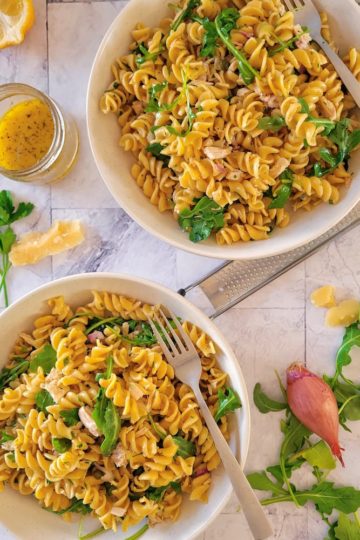 two bowls of pasta salad with tuna, arugula on the top and side with a shallot, a half a lemon and a small jar of the dressing in the background