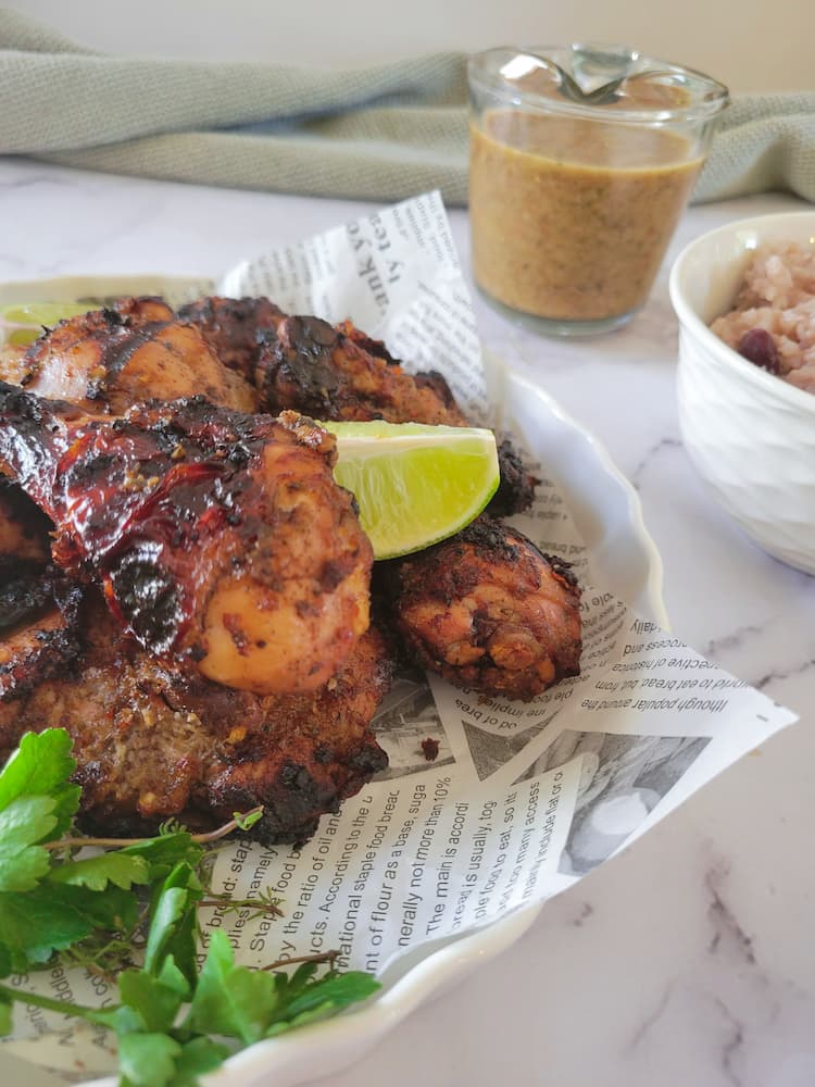 side view of newspaper lined plate with jerk chicken pieces garnished with fresh lime wedges and parsley, bowl of rice and peas and jerk sauce in the background