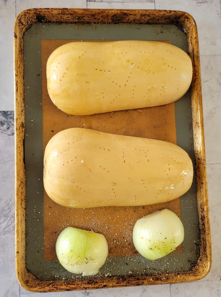 two halves of a butternut squash pierced with a fork face down on a baking sheet with a halved onion