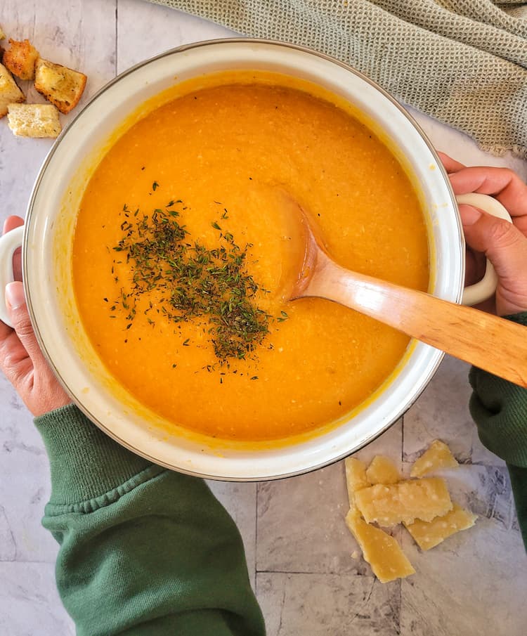 hands holding a pot of butternut squash soup with chopped fresh parsley, parmesan cheese and croutons in the background
