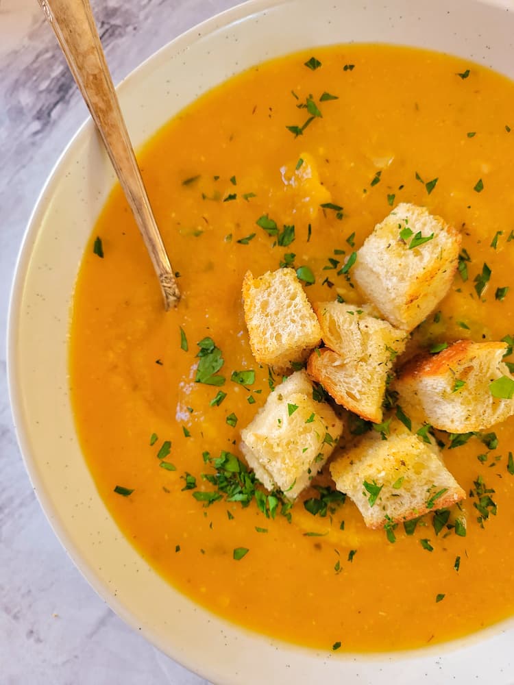close up of a bowl of butternut squash soup with a spoon in it, garnished with croutons and chopped fresh parsley
