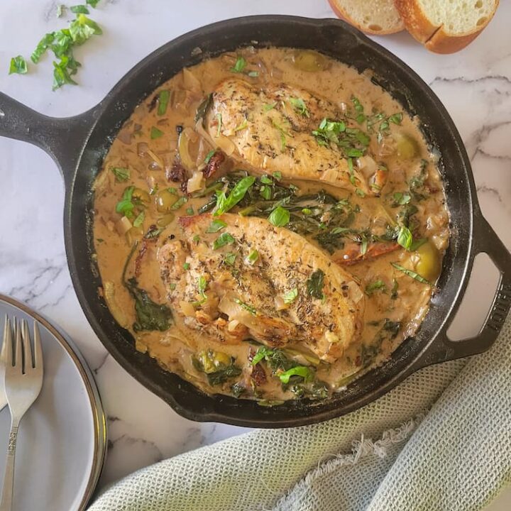 cast iron skillet with 2 chicken breasts in a creamy sauce with fresh herbs and olives, 2 slices of bread in the background, forks/plates on the bottom