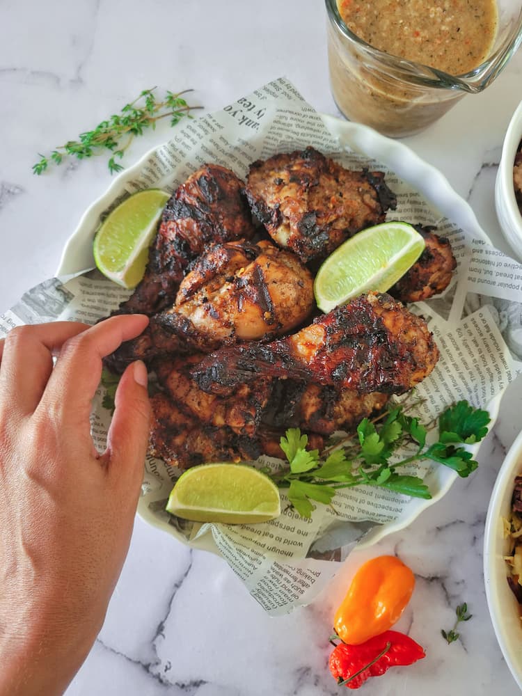 newspaper lined plate with jerk chicken pieces garnished with fresh lime wedges and parsley, jerk sauce and habaneros in the background, hand touching a drumstick on the plate