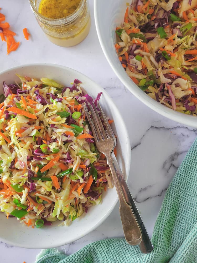 bowl of coleslaw with 2 forks next to another bowl with the rest, jar of dressing next to shredded carrots in the background