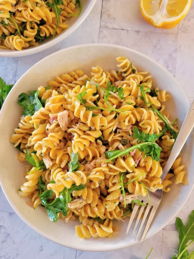 bowl of pasta salad with tuna, topped with arugula, fresh herbs and a half a lemon in the background next to another bowl of pasta salad
