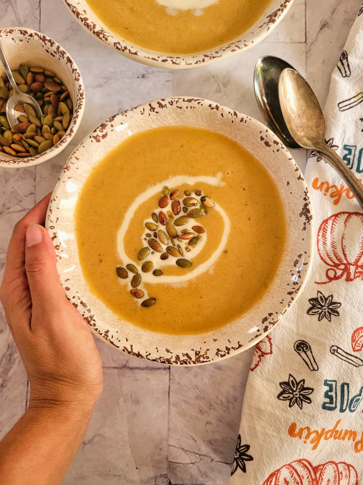 hand holding a bowl of pumpkin soup with a swirl of heavy cream and toasted pepitas, ramekin of more pepitas in the background with two spoons, another bowl of soup in the background