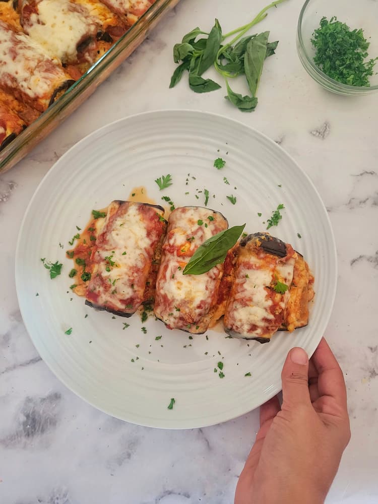 hand holding a plate with three eggplant rollatini next to a baking dish with the rest, chopped parsley and fresh basil in the background and on top