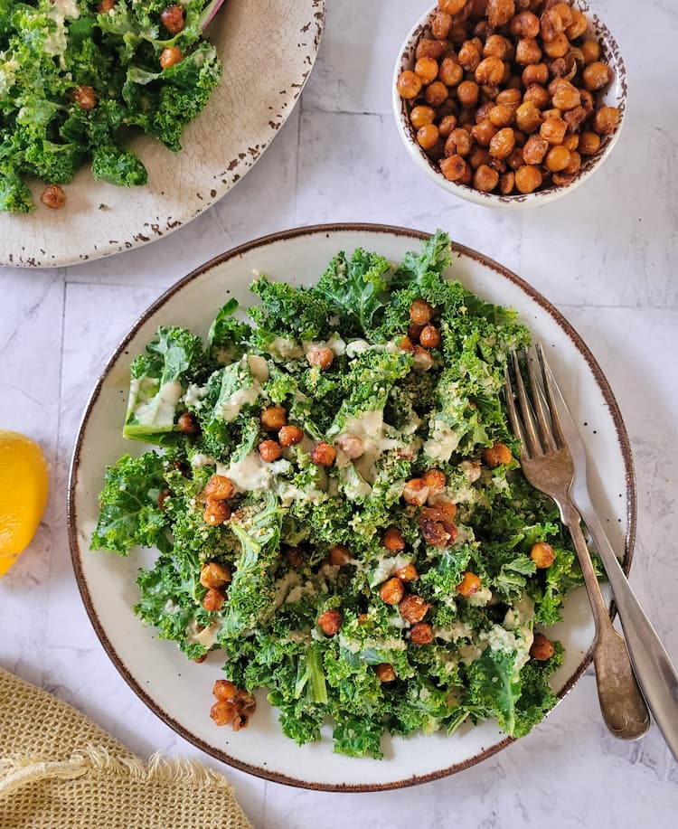 plate of kale caesar salad with roasted chickpeas and a creamy dressing, another plate of salad in the background next to a small bowl of roasted chickpeas and a lemon wedge