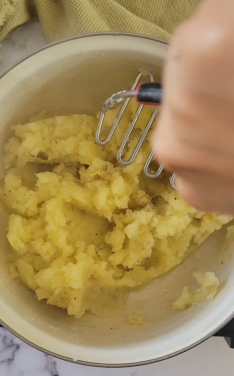 hand with a potato masher mashing a pot of potatoes