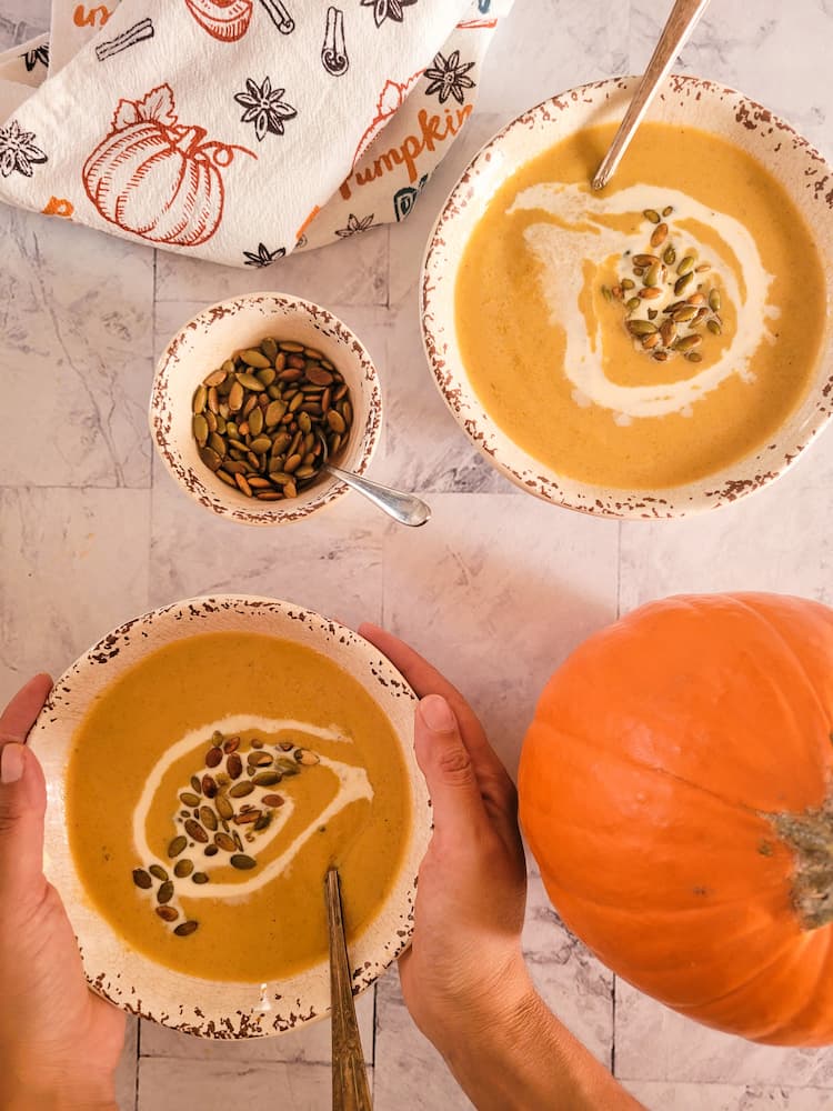 hands wrapped around a bowl of pumpkin soup with toasted pepitas and a swirl of heavy cream, another bowl of soup in the background, sugar pie pumpkin on the bottom right, ramekin of toasted pepitas in the middle