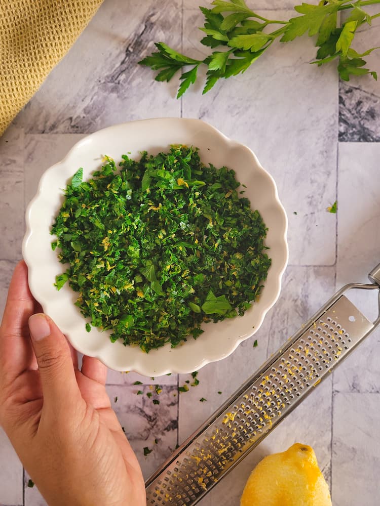 hand holding a bowl of gremolata next to a zested lemon and microplane, some fresh parsley in the background