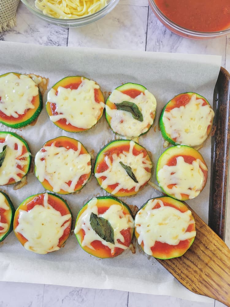 parchment lined baking sheet with cooked zucchini pizza rounds topped with marinara sauce, mozzarella cheese and basil, wooden spatula lifting one up