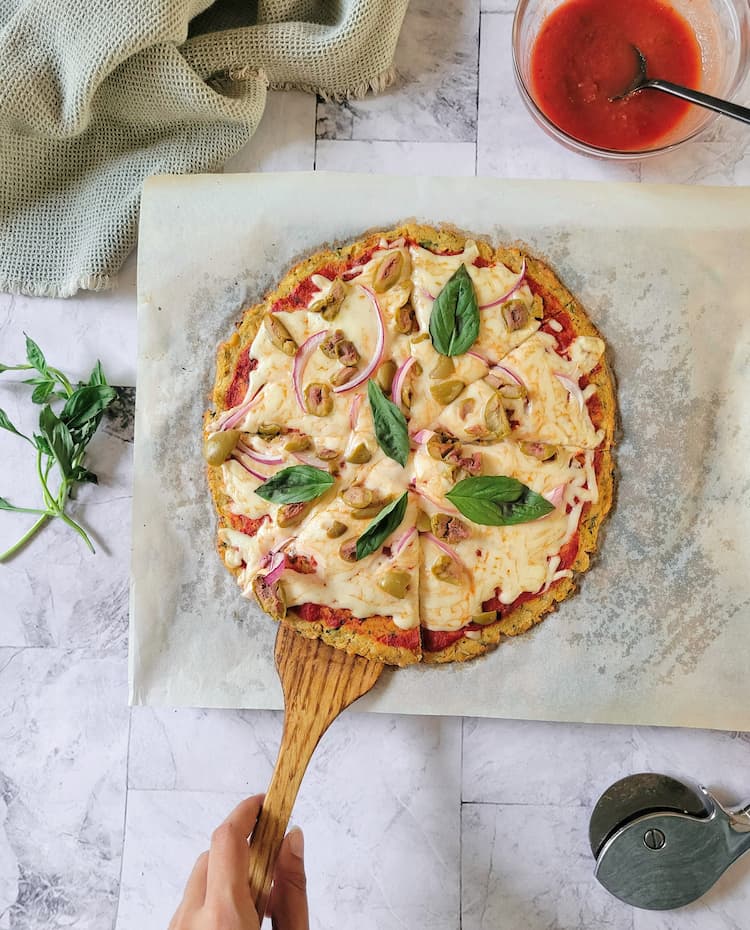 cauliflower crust pizza topped with tomato sauce, mozzarella cheese, fresh basil, green olives and red onion on a parchment lined pizza stone, hand with a wooden spatula lifting up a slice, bowl of tomato sauce, pizza cutter and fresh basil in the background
