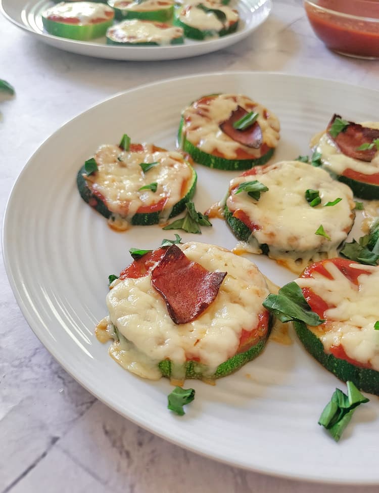 side view of zucchini pizza bites with mozzarella cheese, pepperoni and chopped fresh basil, another plate with more in the background