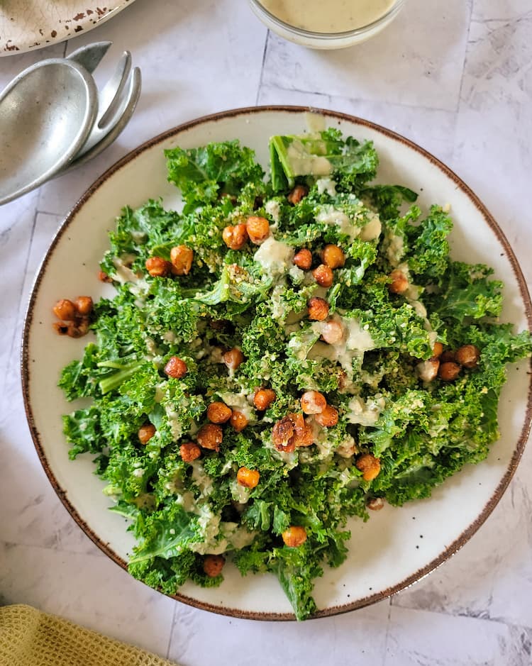 plate of kale caesar salad with roasted chickpeas and a creamy dressing, silver salad tongs in the background next to a cut off bowl of dressing