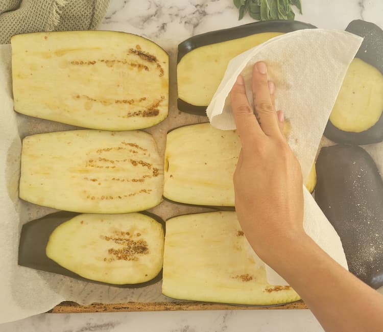 hand with a paper towel patting down sliced eggplant on a parchment lined baking sheet