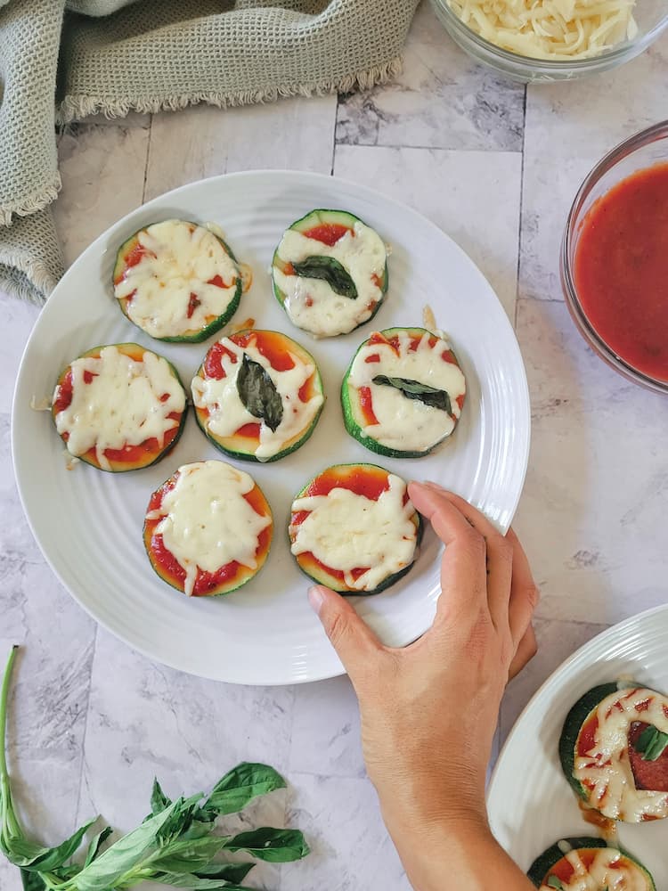 hand grabbing a zucchini pizza bite off a plate with several, marinara, fresh basil, mozzarella cheese and more bites in the background