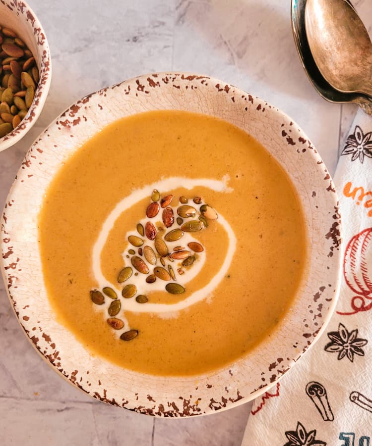 bowl of pumpkin soup with a swirl of heavy cream and toasted pepitas, two spoons in the background with a ramekin of more pepitas