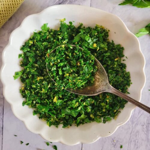 bowl of gremolata with a spoon in it, fresh parsley and a zested lemon in the background