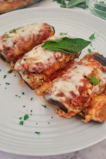 side view of 3 eggplant rollatini on a plate garnished with chopped parsley and a basil leaf