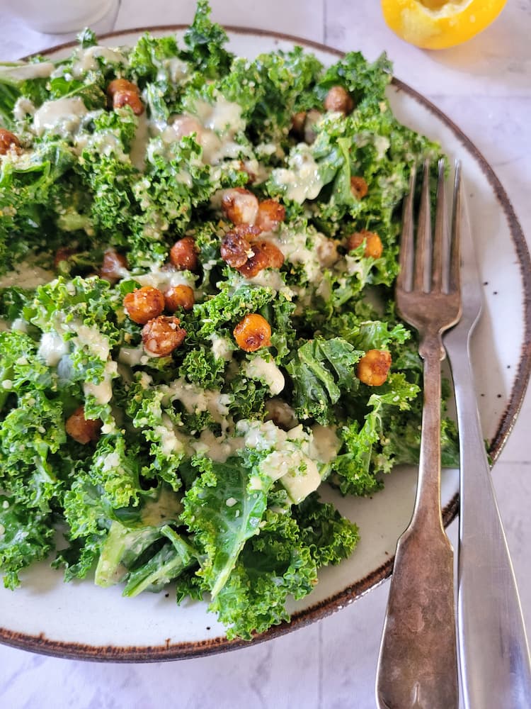 plate of kale caesar salad with roasted chickpeas and a creamy dressing, lemon in the background, 2 forks on the plate
