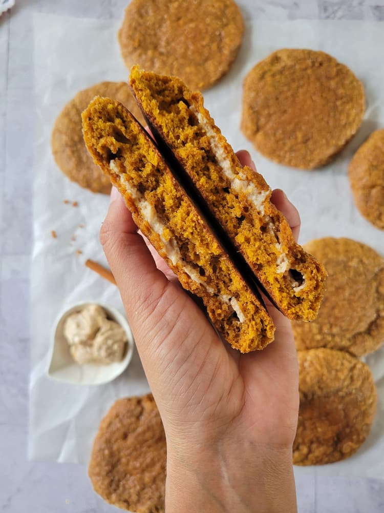 hand holding a pumpkin spice cookie split in half showcasing a thin cream cheese filling, other cookies in the background