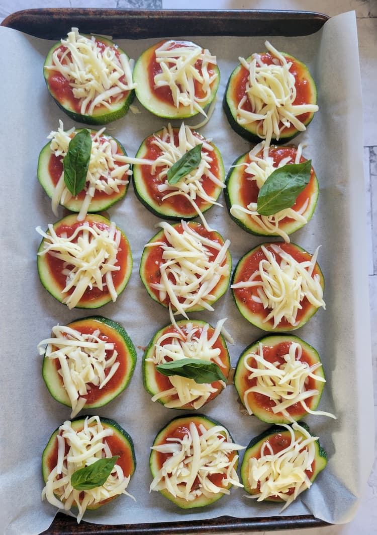 parchment lined baking sheet with uncooked zucchini pizza bites, topped with marinara sauce, shredded cheese and fresh basil