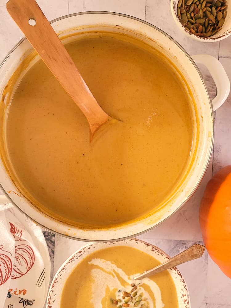 pot of pumpkin soup with a wooden spoon next to a ramekin of toasted pepitas, a sugar pie pumpkin and a bowl of the soup with a heavy cream swirl and pepitas
