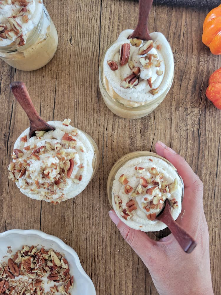 4 jars of pumpkin mousse topped with pecans and whipped cream, wooden spoon in each jar, hand grabbing one of the jars, pumpkins and bowl of chopped pecans in the background