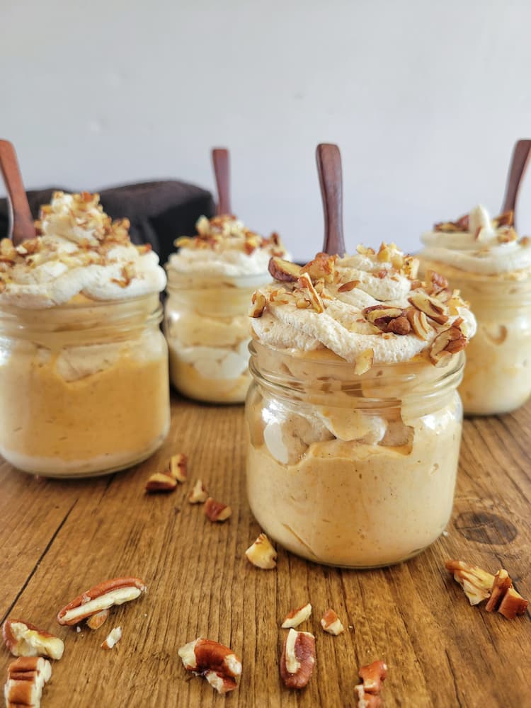 4 jars of pumpkin mousse topped with pecans and whipped cream, chopped pecans in the front, wooden spoon in each jar