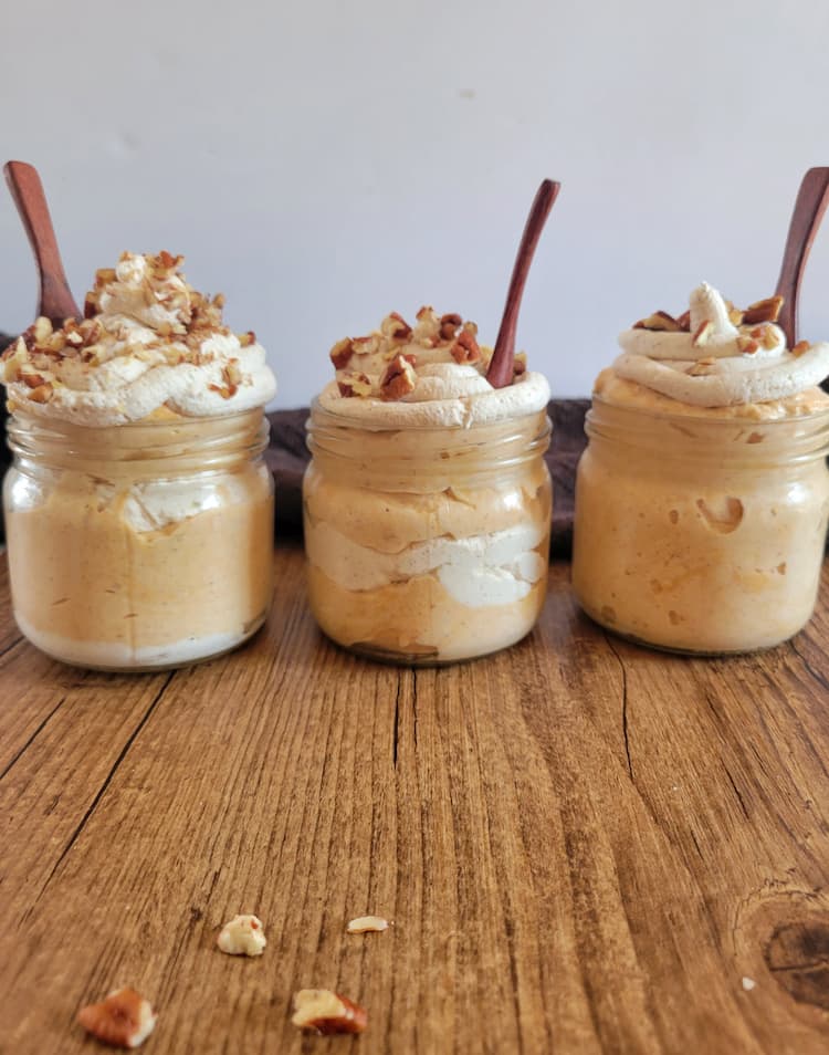 three jars of pumpkin mousse, layered with whipped cream, topped with pecan halves which are also in the background, each jar has a small wooden spoon