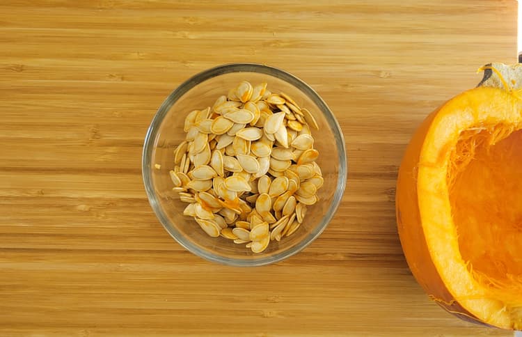 a bowl of pumpkin seeds next to a half cored pumpkin