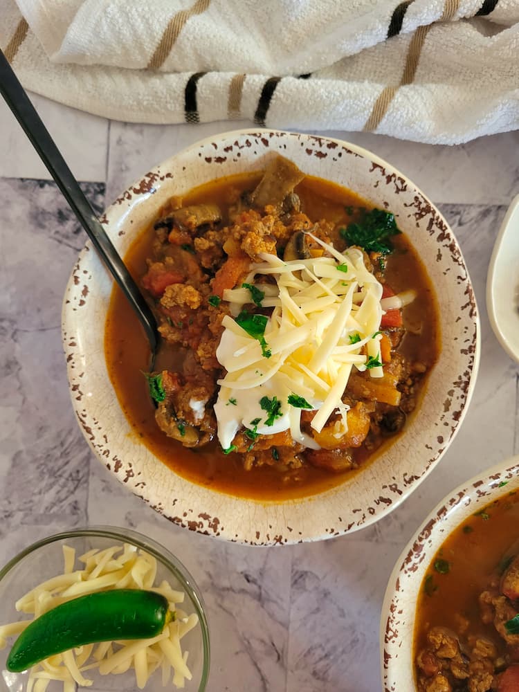 bowl of pumpkin chili garnished with grated mozzarella, a dollop of sour cream and fresh chopped parsley, spoon in the bowl, ramekin of grated mozzarella with a whole jalapeno next to another bowl of chili in the background