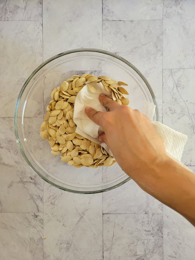 hand with a paper towel patting down some whole raw pumpkin seeds in a bowl