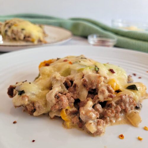 side view of a piece of a philly cheesesteak casserole on a plate, another plate with another piece in the background with a ramekin of chili flakes