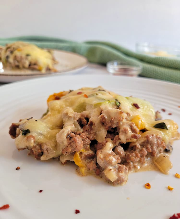 side view of a piece of a philly cheesesteak casserole on a plate, another plate with another piece in the background with a ramekin of chili flakes