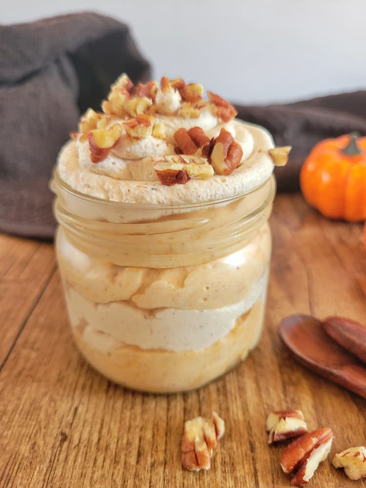 jar of pumpkin mousse, layered with whipped cream, topped with pecans, pecans, spoons and a small pumpkin in the background