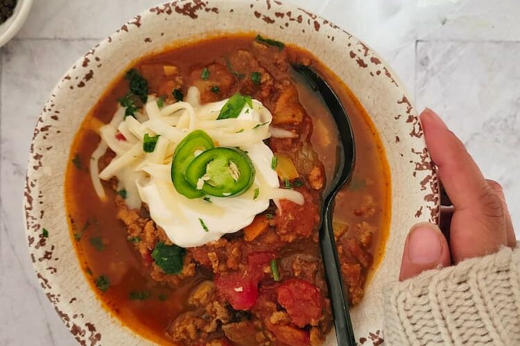 hand on a bowl of pumpkin chili with a spoon, topped with sour cream, sliced jalapeno, shredded mozzarella cheese, bowl of more cheese in the bottom background