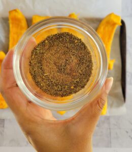 hand holding a ramekin of spices over a baking sheet of pumpkin wedges