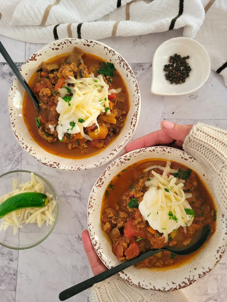 two bowls of pumpkin chili garnished with grated mozzarella, a dollop of sour cream and fresh chopped parsley, hands hugging one bowl, spoons in each bowl, ramekin of peppercorns and mozzarella with a whole jalapeno in the background
