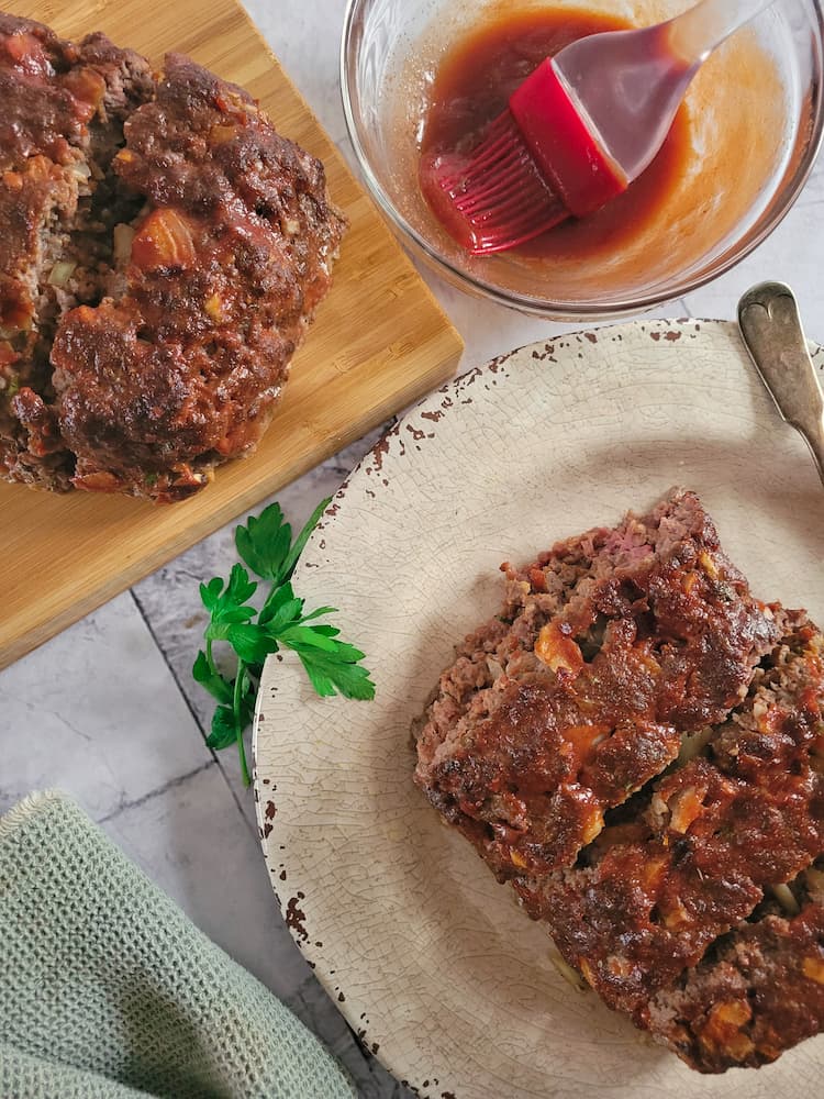 a plate with sliced meatloaf, next to a cutting board with more meatloaf and a bowl of bbq sauce with a basting brush